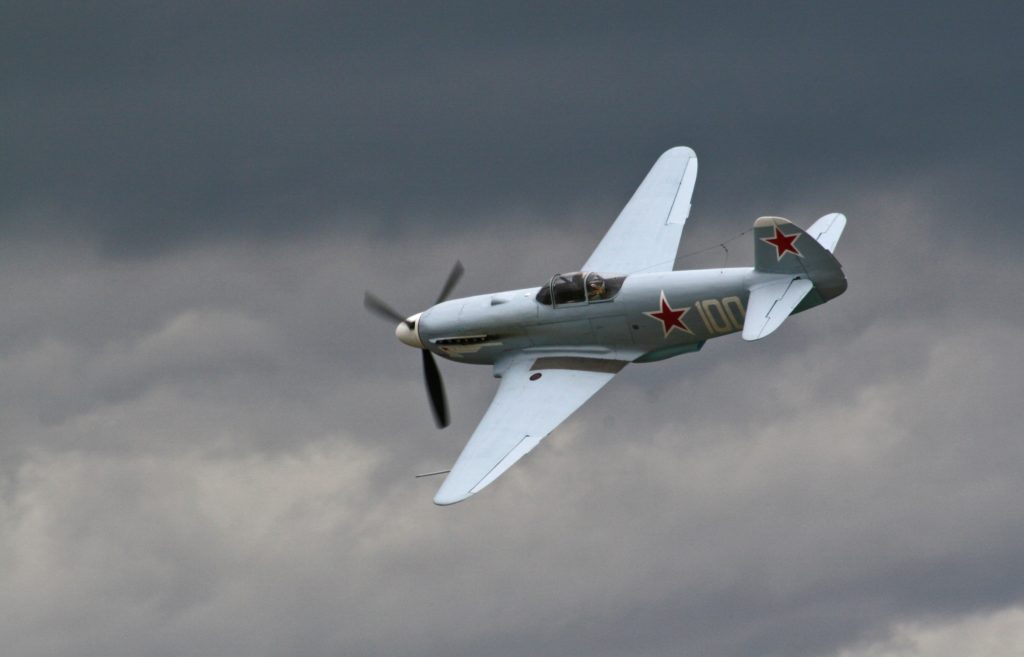 Gray biplane flies through cloudy skies with a red star and the number 100 emblazoned on the side