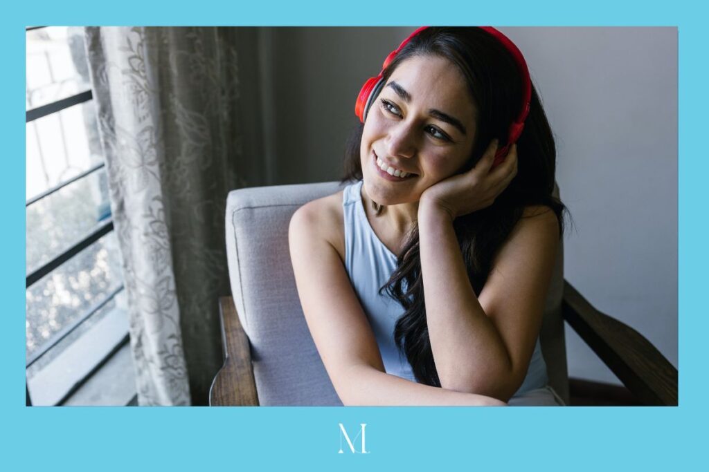 Woman with long dark hair wearing red headphones looks to her right out an open window. She's smiling and pressing one hand to her left ear over the headphones. The border is turquoise and thicker on the bottom, where there's a white stylized "M" in the center.