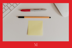 Empty white desk with a red Sharpie, black marker, and empty yellow post-it note lined up. Surrounded by a red border with a stylized white "M" logo on the bottom