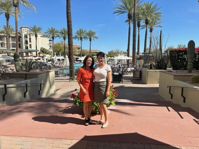 Charlene wears an orange dress and Eva a white top and green skirt. They stand arm-in-arm on a pool deck surrounded by blue sky and palm trees