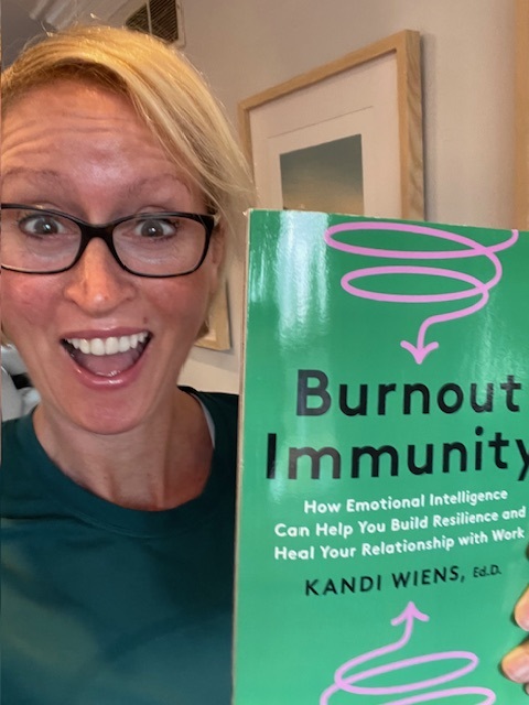 Kandi Wiens wears glasses and an excited smile, and holds up her book "Burnout Immunity"