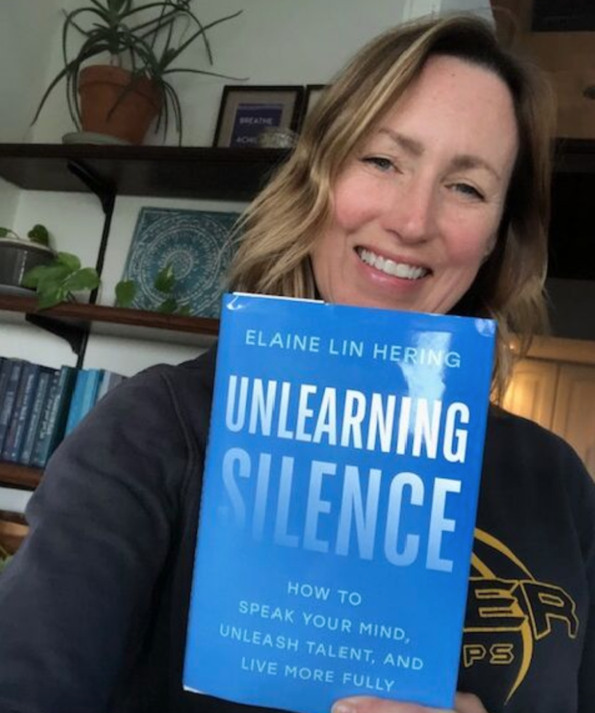 Shonda Moralis is a white woman who smiles and holds the book "Unlearning Silence" by Elaine Lin Hering