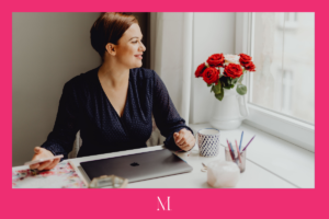 Woman in professional dress sits with a laptop and phone, and smiles out the window